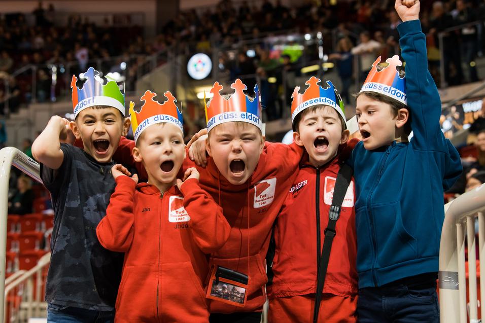 Gruppe von fünf jubelnden Kindern mit farbigen Kronen und roten Sweatshirts in einer Sportarena.