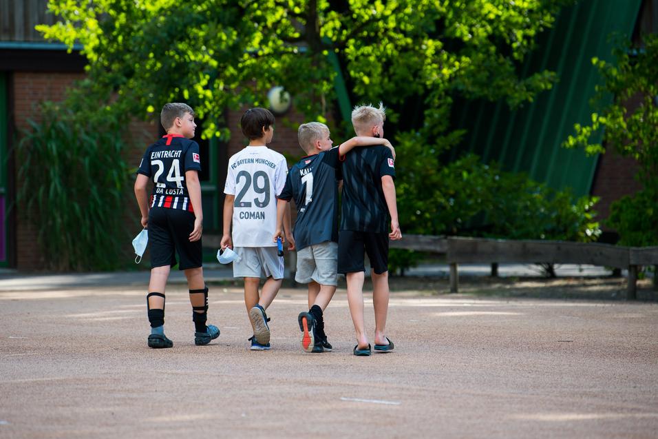 Vier Kinder in Fußballtrikots gehen Arm in Arm über einen Platz, umgeben von Bäumen und einer ruhigen Atmosphäre.